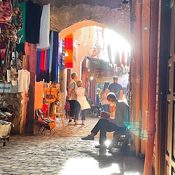 Ein älterer Mann liest für sich in der Medina in Marrakesch während um ihn herum Menschen die Waren der kleinen Läden anschauen. 