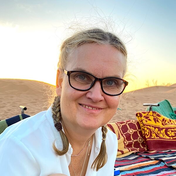 Jutta Büttner lächelt und sitzt auf Berberteppichen in der Wüste. 