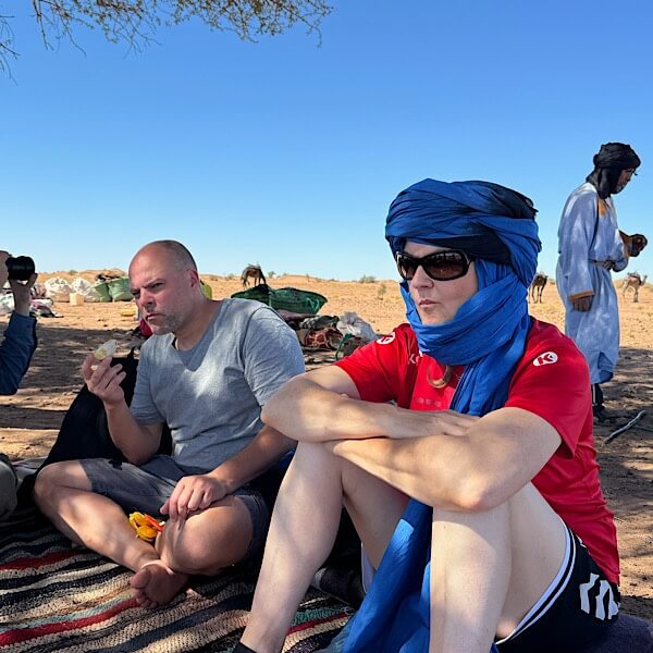 Mann und Frau sitzen in der Wüste. Im Hintergrund Kamele. 