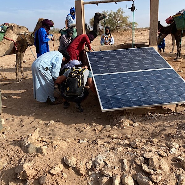 Eine betonierte Wasserstelle in der Wüste mit großem Photovoltaik-Panel im Vordergrund. Menschen sitzen am Boden und versuchen die Elektrik zu reparieren. 