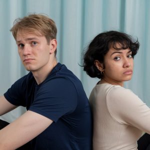 Ein Mann und eine Frau sitzen Rücken an Rücken und schauen frustriert in die Kamera.