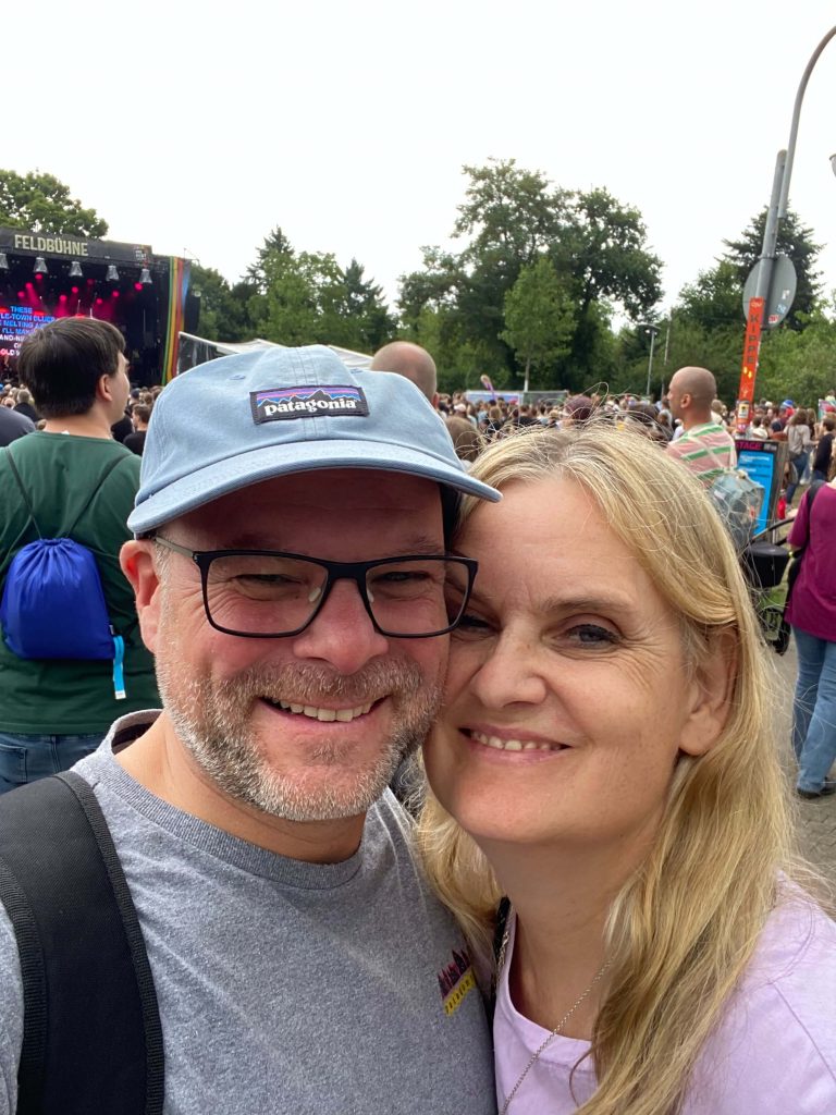 Frau schmust mit Mann. Beide schauen in die Kamera. Es sind Menschen im Hintergrund.