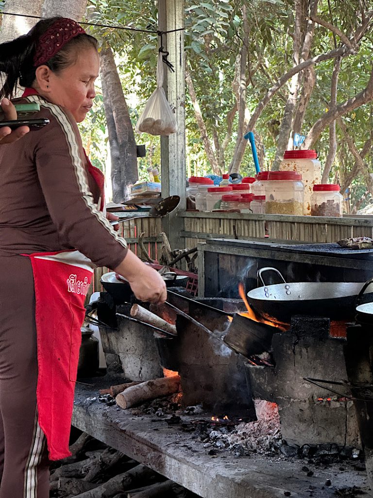Eine Frau steht vor einer traditionellen kambodschanischen Kochstelle in einem Pavillion in Kompong Thom. Unter der Pfanne ist das Holzfeuer zu sehen
