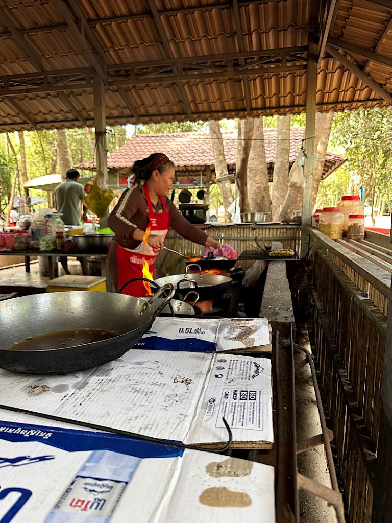 Eine Frau steht in einem Pavillion. Sie kocht. Im Vordergrund sind Pfannen zu sehen. Kompong Thom.