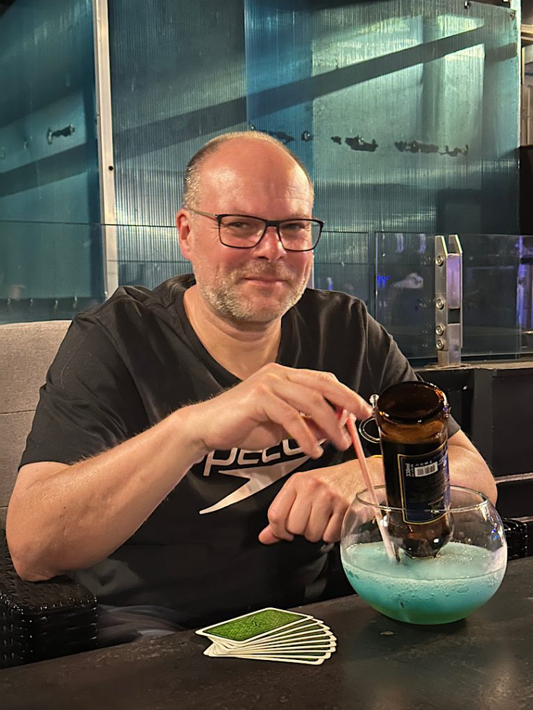 Ein Mann sitzt am Tisch. Vor ihm ein Hanuman-Cocktail. Eine Flasche Bier steckt kopfüber in einem Cocktailglas. Das Bier läuft langsam in den Margaritha