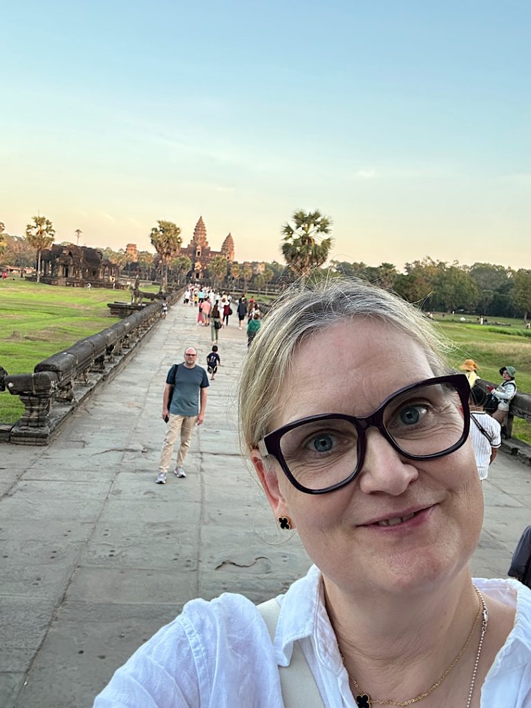 Frau macht Selfie mit Tempel Angkor Wat im Hintergrund. Die Sonne geht unter und färbt den Tempel golden ein.