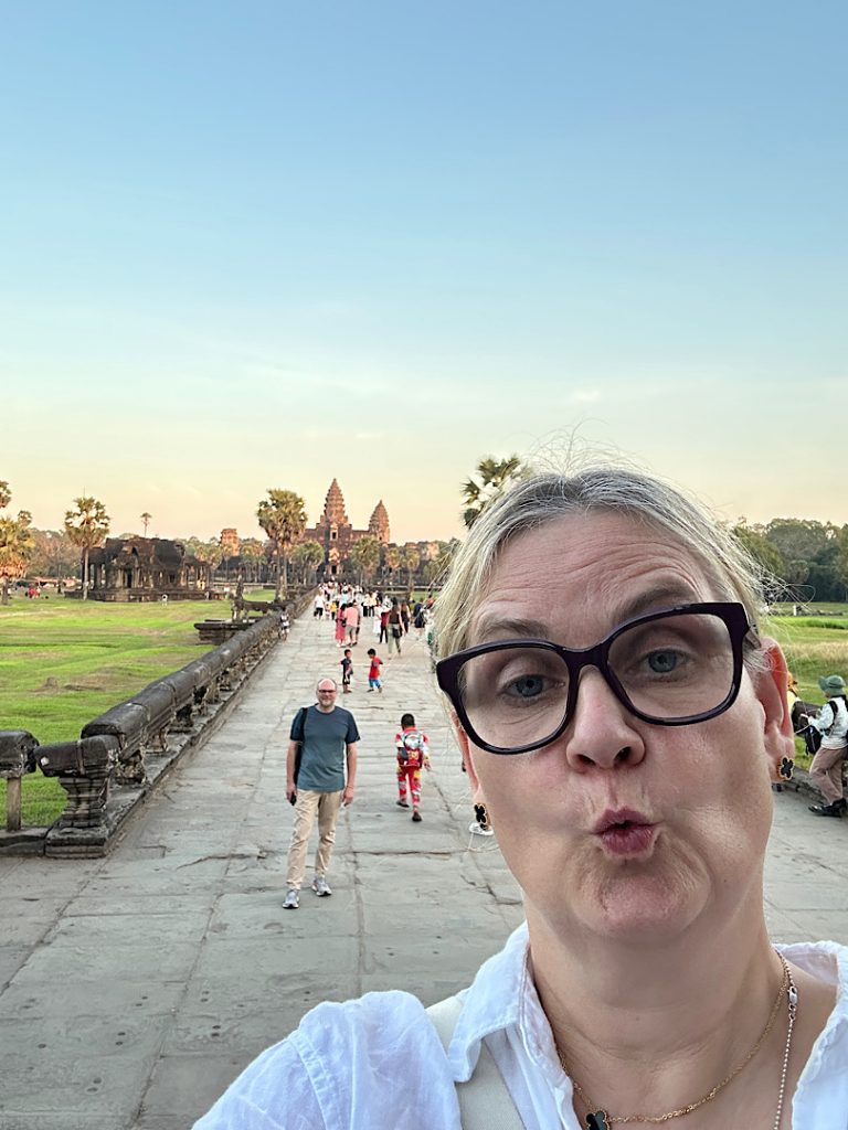 Frau macht Selfie mit Tempel Angkor Wat im Hintergrund. Die Sonne geht unter und färbt den Tempel golden ein.