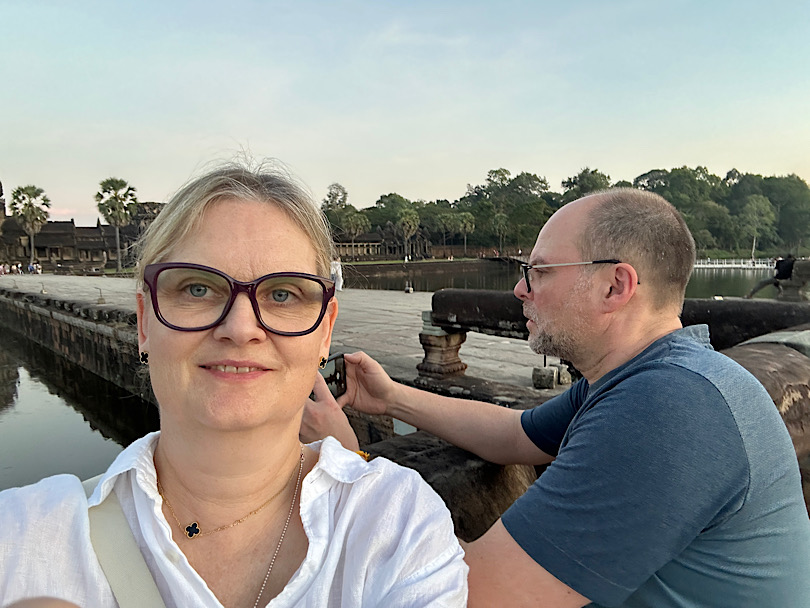 Frau macht Selfie. Im Hintergrund steht ein Mann seitlich, der ein Motiv außerhalb des Bildes fotografiert.