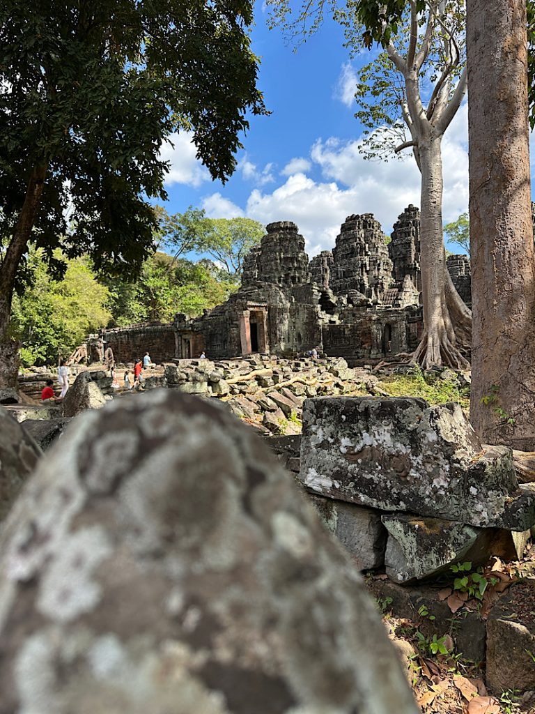Im Vordergrund ist ein Stück Sandstein. Dahinter ragt ein Khmer-Tempel auf. Darüber sieht mn einen blauen Himmel