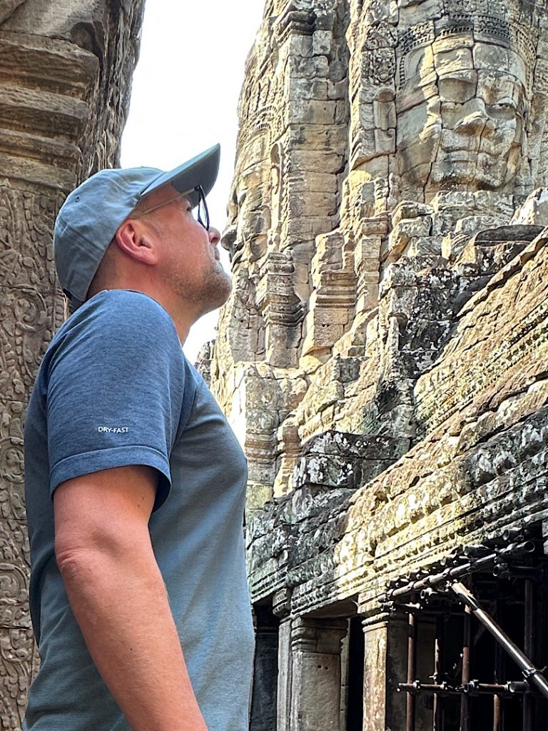 Ein Mann steht seitlich zur Kamera. Im Hintergrund ist ein Tempel von Angkor Thom zu sehen. Durch die Perspektive entsteht der Eindruck, dass er mit seiner Nase, die Nase des Steingesichts berührt.