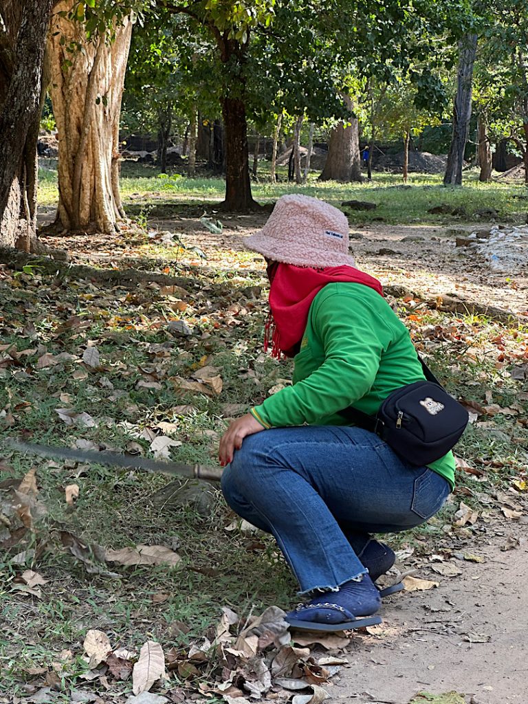 Im Vordergrund ist eine Frau zu sehen. Sie trägt eine blaue Hose, ein grünes Tshirt, einen Fischerhut und ein rotes Halstuch. Sie hockt auf den Fersen und schwingt ein Messer. Sie schneidet den Rasen.