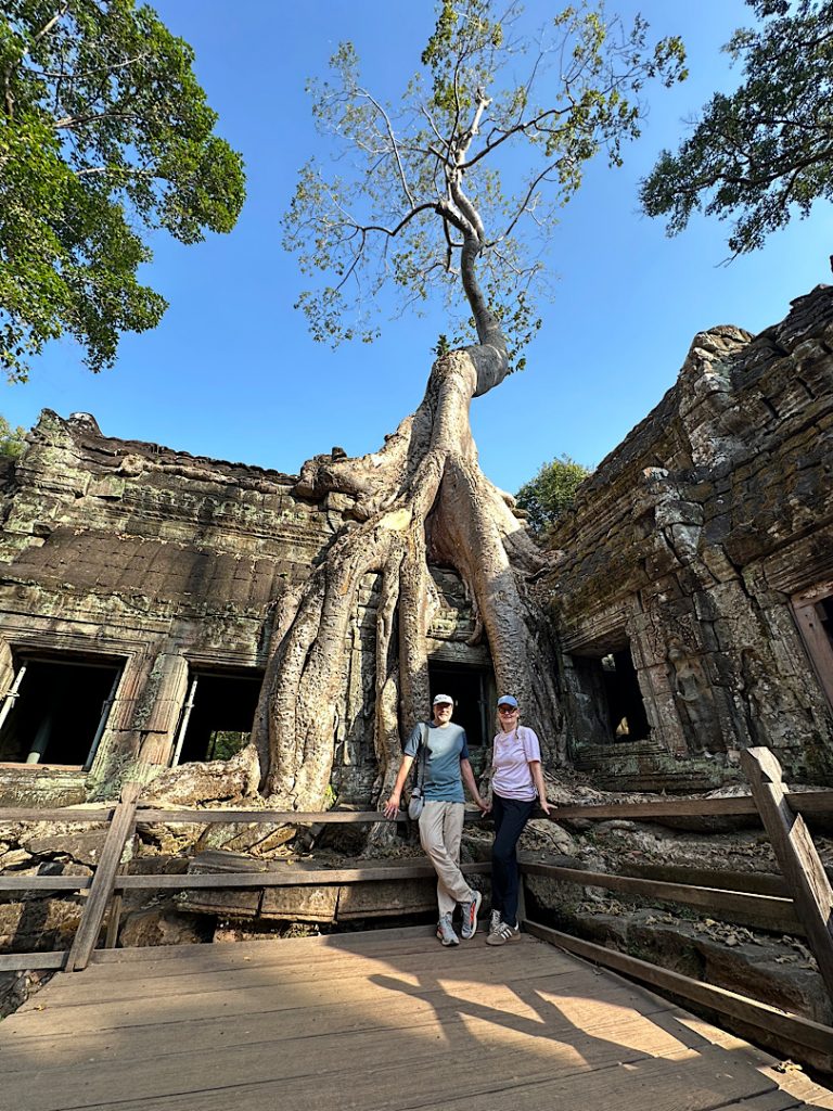 Zwei Menschen stehen vor einem riesigen Baum, der seine Wurzeln über einen kambodschanischen Tempel gelegt hat.
