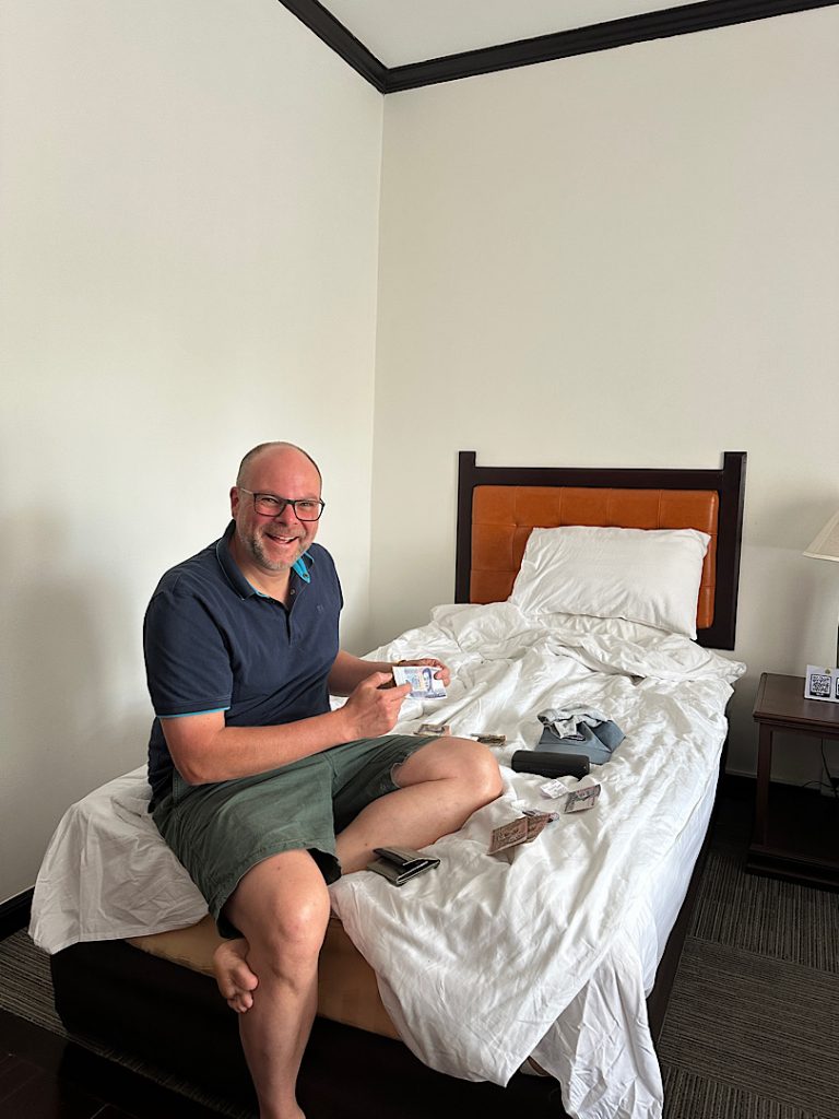 Mann sitzt auf einem Bett in einem Hotelzimmer. Vor ihm liegen Geldscheine. Er lacht in die Kamera und zeigt einen Schein.