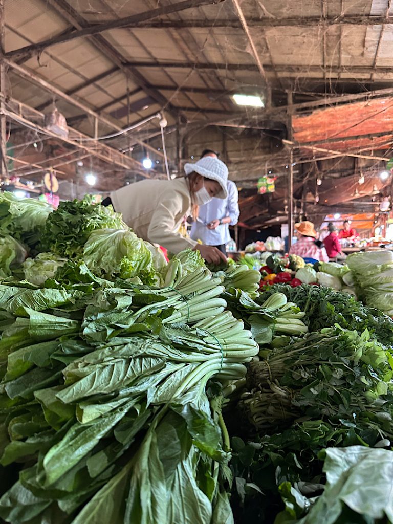 Hinter einem Berg von grünem Gemüse wie Wasserspinat ist eine Frau zu sehen, die einen Hut trägt. Sie ist die Verkäuferin.
