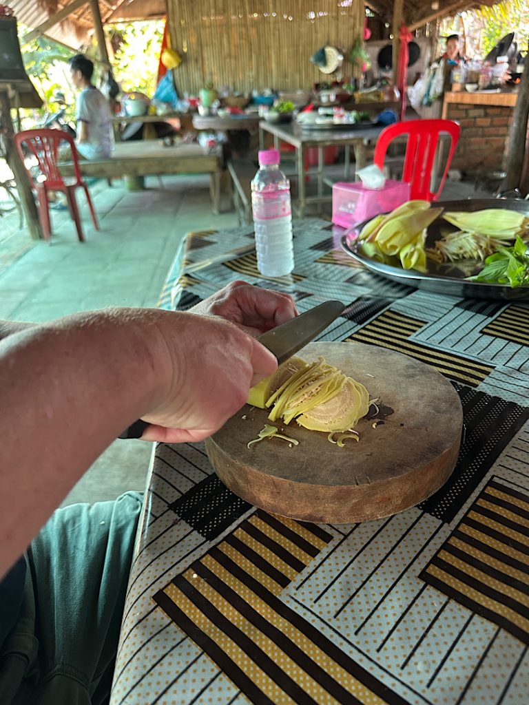 Auf einem Holzbrett liegt eine halbe Bananenblüte. Sie wird in feine Streifen geschnitten von zwei Händen.