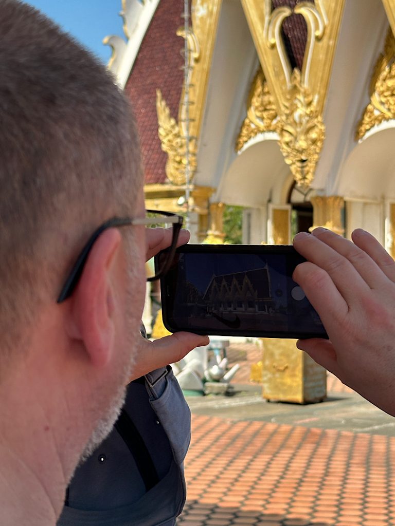 Über seine Schulter fotografiert ist der halbe Kopf eines Mannes und sein Ohr zu sehen. Er hält sein Handy zum Fotografieren bereit.