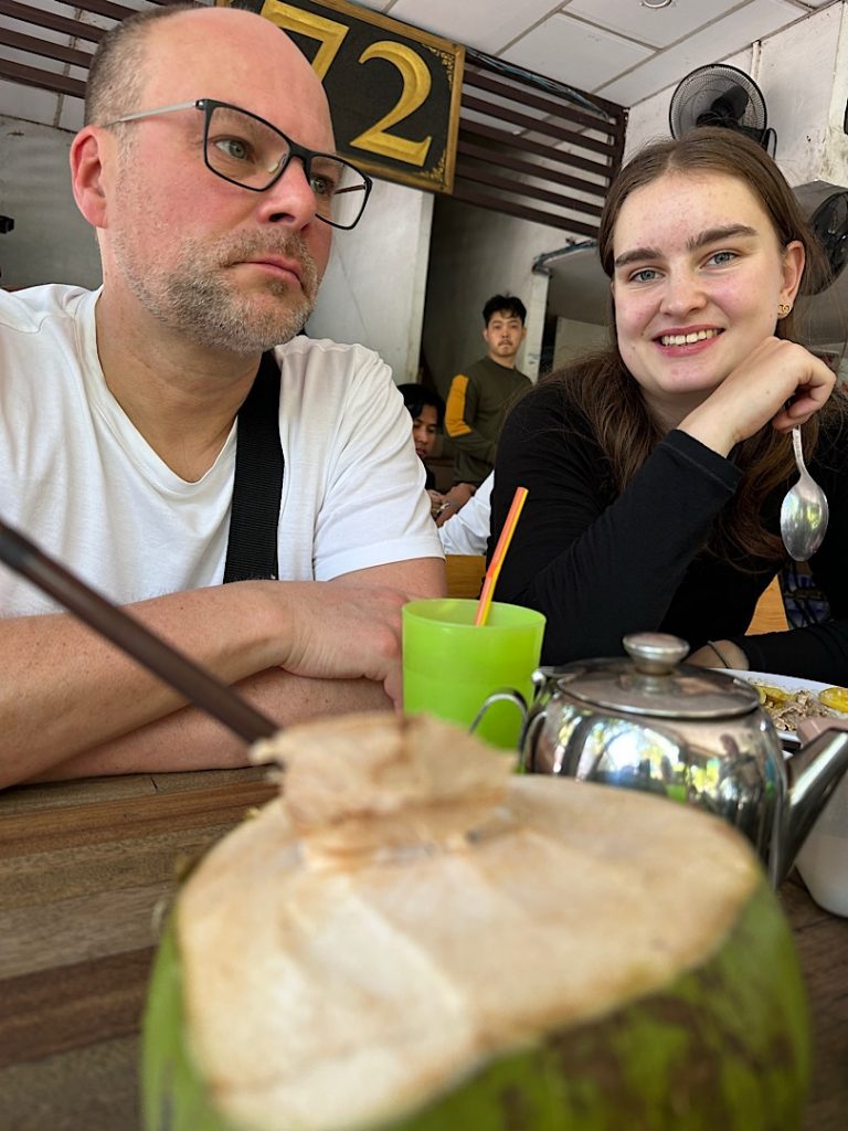 Ein Mann und eine Frau sitzen an einem Tisch in Phnom Penh, Kambodscha. Im Vordergrund eine Kokosnuss zum Trinken, eine Teekanne und ein grüner Becher.