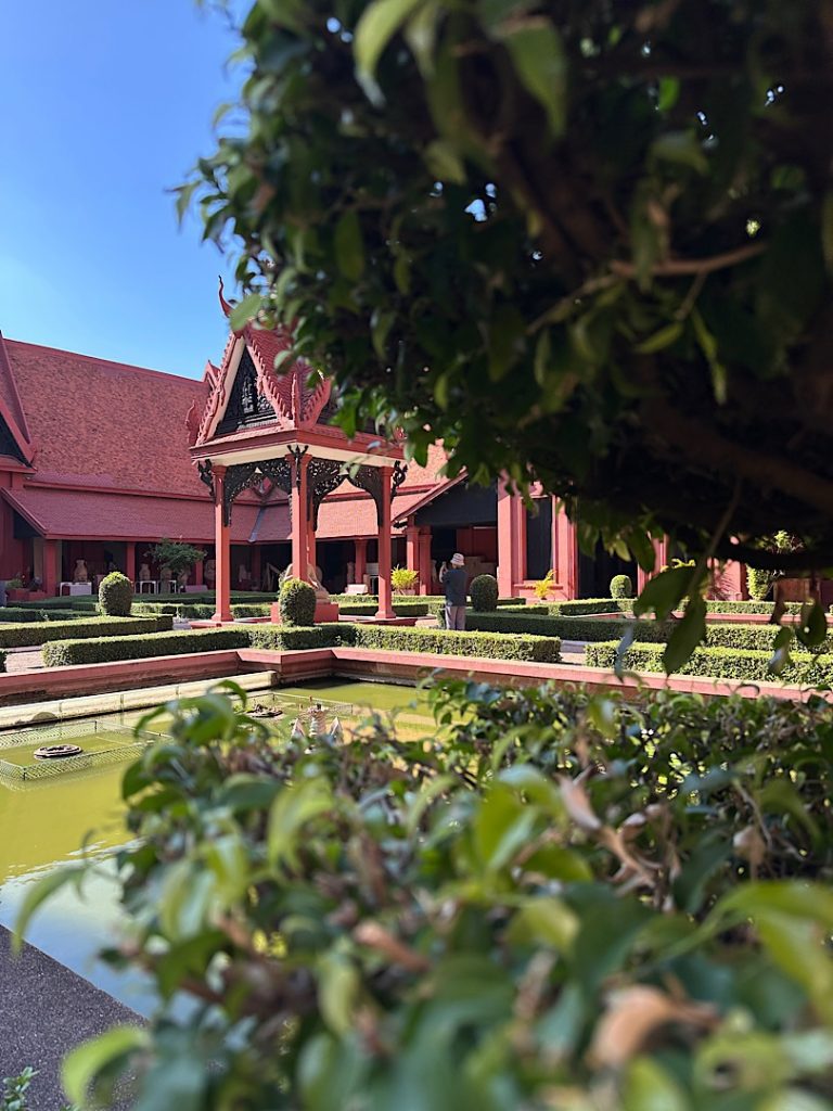 Zwischen einem Baum hindurch ist das Gebäude des Nationalmuseums in Phnom Penh, Kambodscha zu sehen. Es ist rot und in Khmerarchitektur.