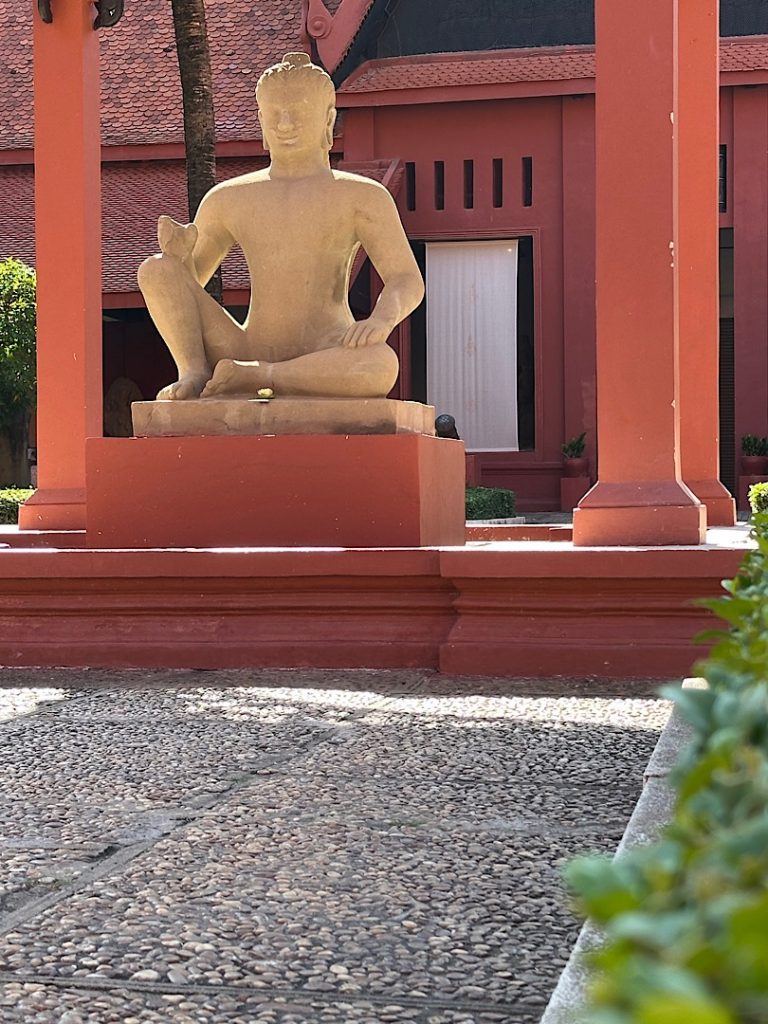 Eine steinerne Buddha-Statue im Nationalmuseum Kambodscha in Phnom Penh. Die Statue steht in einem kleinen, rot gestrichenen Pavillon.