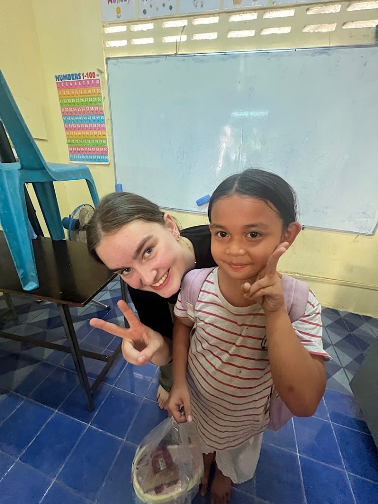 Ein kambodschanisches Kind lächelt in die Kamera. Dahinter bückt sich eine Frau und umarmt das Kind leicht. Sie zeigt ein Viktory-Zeichen mit der rechten Hand.