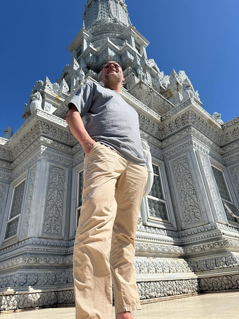 Mann posiert vor einer Pagode. Er trägt ein graues Shirt und eine beige Hose. Er schaut über die Kamera hinweg.