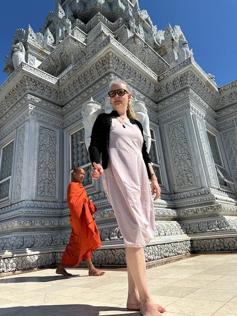 Frau steht vor einer Pagode für ein Selfie. Sie träg ein rosa Kleid und eine schwarze Strickjacke. Hinter ihr läuft ein buddistischer Mönch durch das Bild.