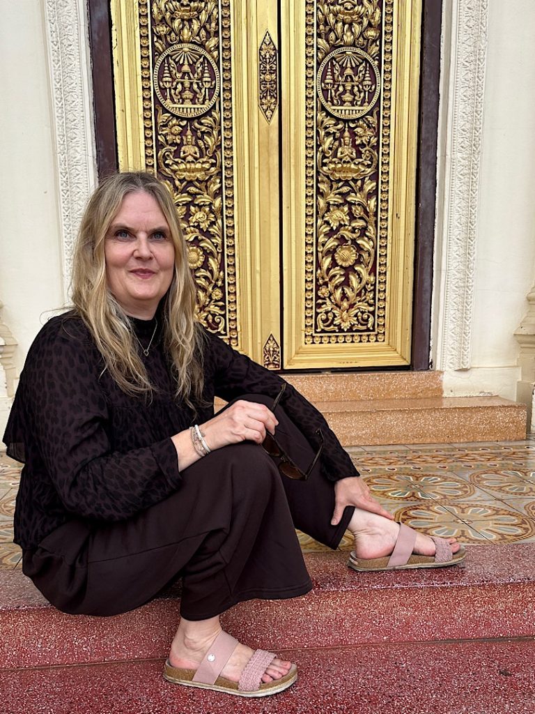 Eine Frau in brauner Kleidung sitzt auf einer roten Treppe vor einer golden verzierten Tür.