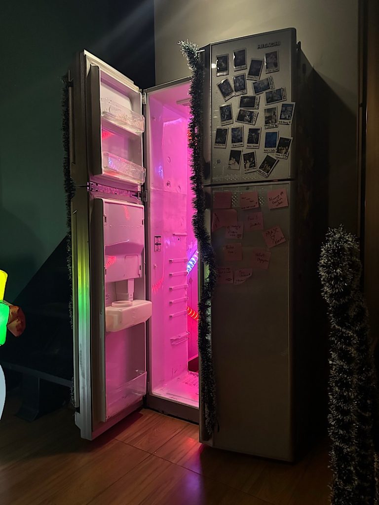 Es ist ein halb geöffneter Kühlschrank zu sehen. Dieser ist die Tür zu einer Bar.