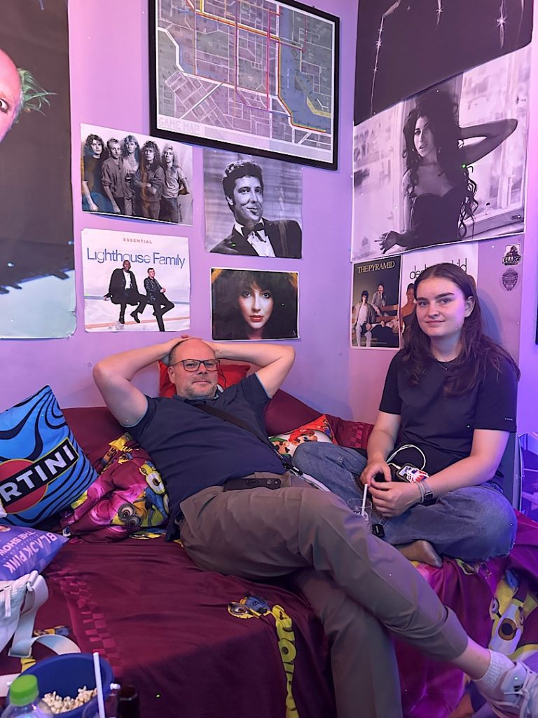 Ein Mann liegt auf einem Bettsofa. Daneben sitzt eine junge Frau. An der Wand hängen Bilder von Sängerinnen und Bands.