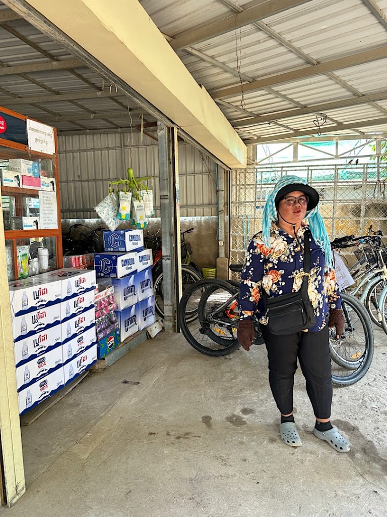 Eine Kambodschanerin, geschützt gegen die Sonne mit Hut, Nackentuch und Handschuhen, steht vor einem Motorrad und einem Regal in einer Halle.