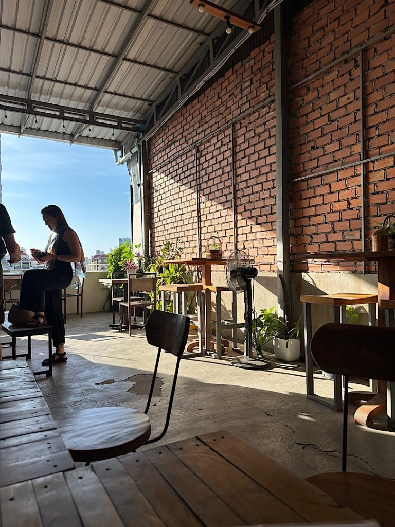 Blick über einen Tisch auf eine Ziegelmauer. Links eine Frau beim Bezahlen. Hinter ihr der Blick über den Russia Market in Phnom Penh Kambodscha