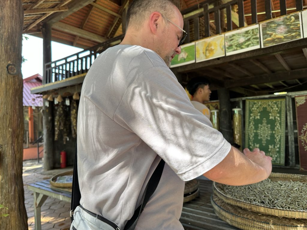 Ein Mann steht an einem Tisch. Er ist von hinten zu sehen. Vor ihm ist ein Korb mit Seidenraupen.