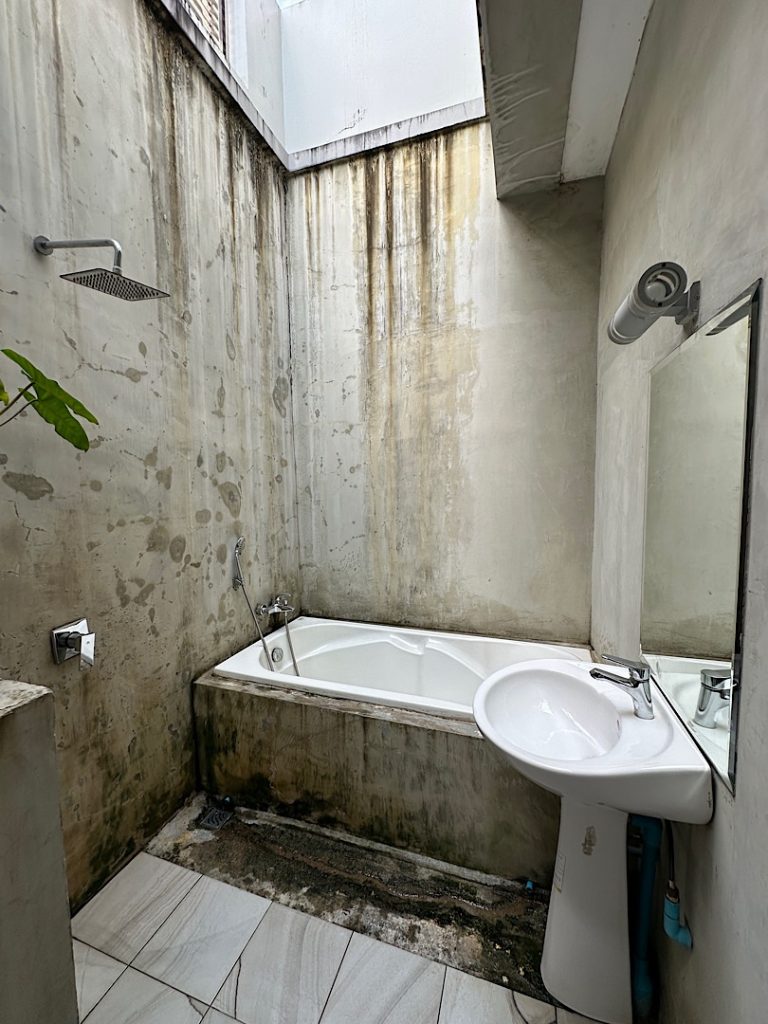 Eine Badewanne ist hinten quer. Davor links ist ein Duschkopf und rechts ein Waschbecken zu sehen.