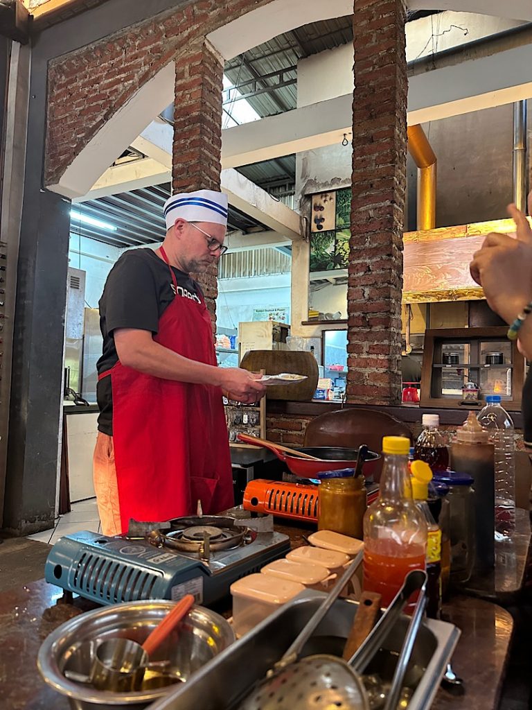 Ein Mann mit roter Schürze und weißer Kochmütze vor einer Gaskochplatte. Auf der Kochplatte ist eine Pfanne.