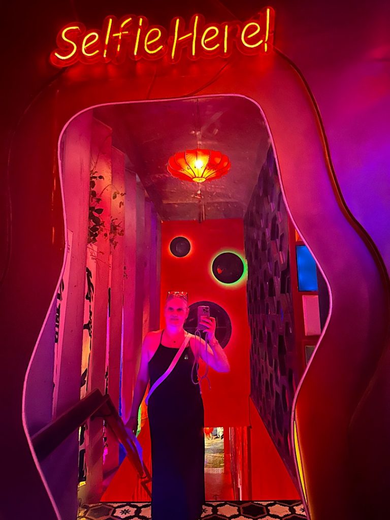 Eine Frau steht vor einem großen Spielgel. Sie fotografiert sich selbst. Das Licht ist künstlich rot.