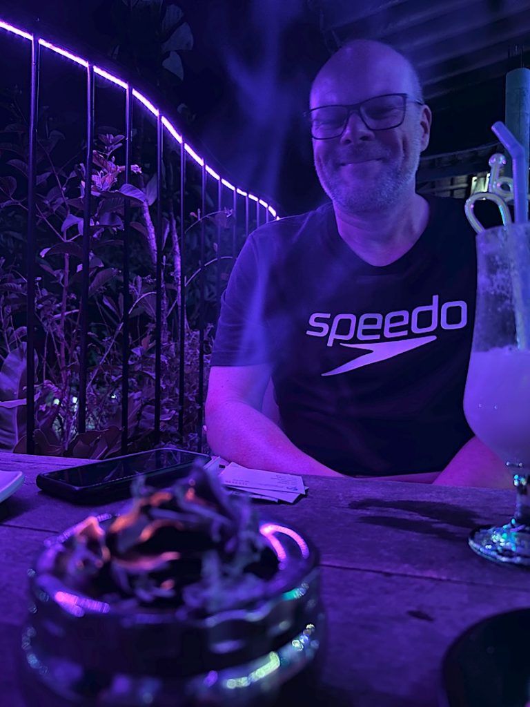 Ein Mann lächelt in die Kamera. Das Licht ist bläulich. Es ist Nacht.