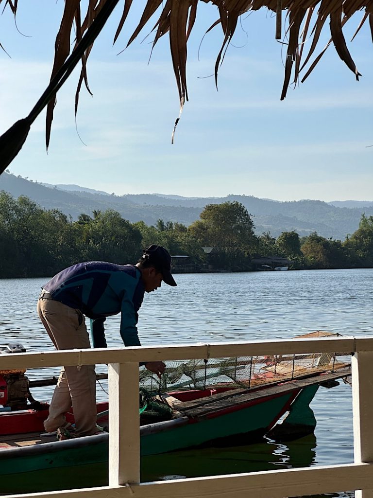 Von Land aus fotografiert ist ein Mann zu sehen, der auf einem Boot steht. Er bückt sich zu seiner Fischreuse, die er ins Wasser lassen möchte.