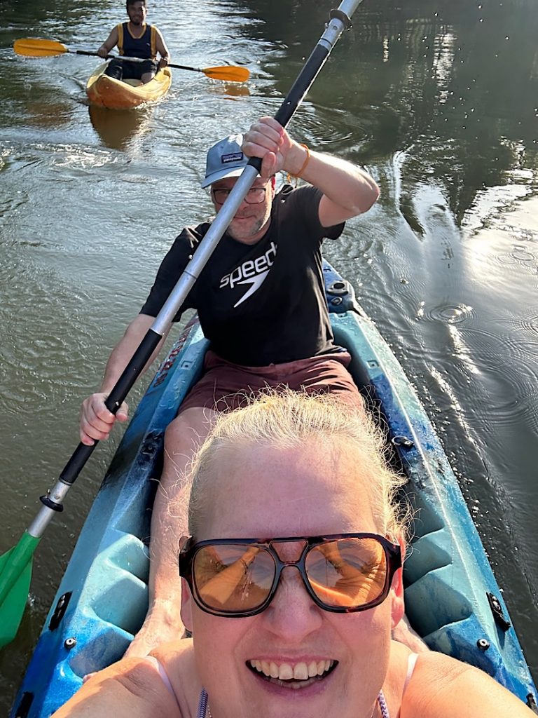 Eine Frau fotografiert über ihren Kopf nach hinten den Mann im Kanu, der rudert. Dahinter ist ein weiteres Kanu mit Mann zu sehen