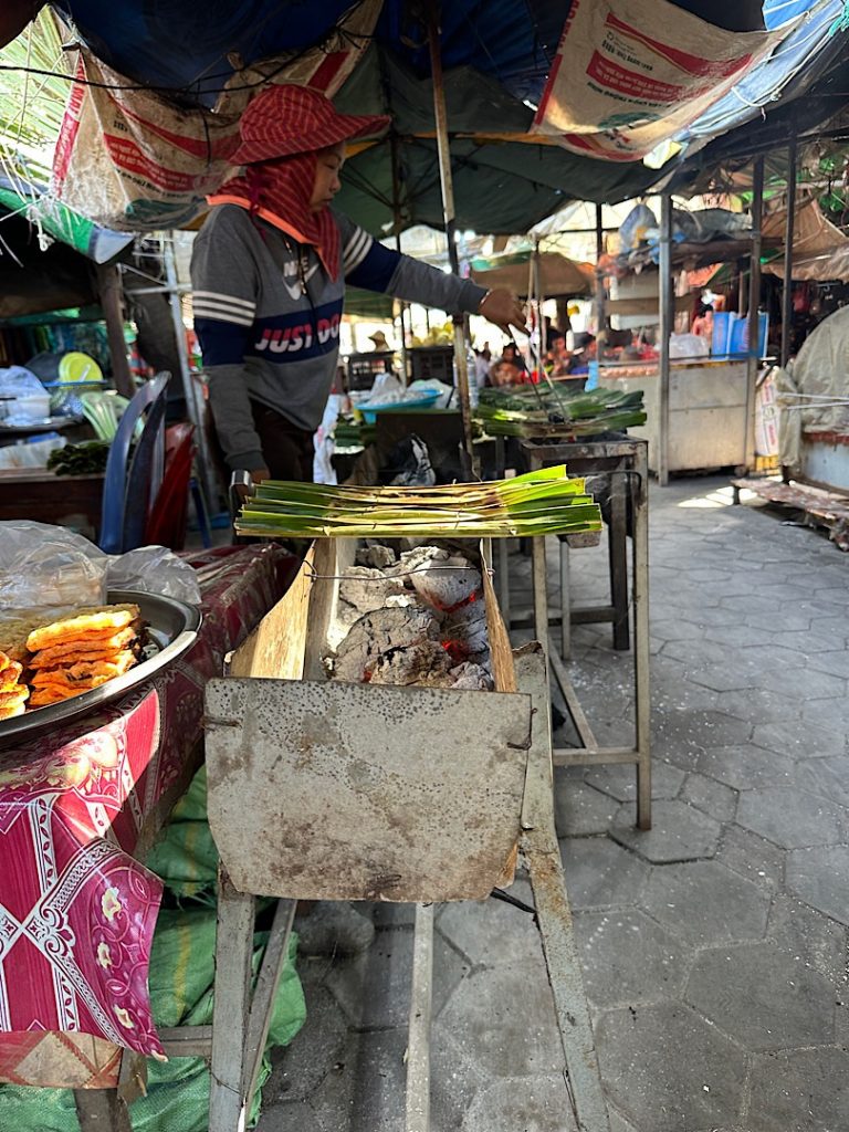 Es ist eine Rinne zu sehen, in der glühende Kohlen lieben. Darauf sind Bananenblätter gelegt. Es ist auf einem Markt in Kep, Kambodscha