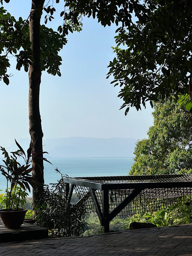 Über ein Tagesbett hinweg ist das Meer fotografiert. Links ist eine Palme. Rechts ein Gebüsch und ein Baum zu sehen.