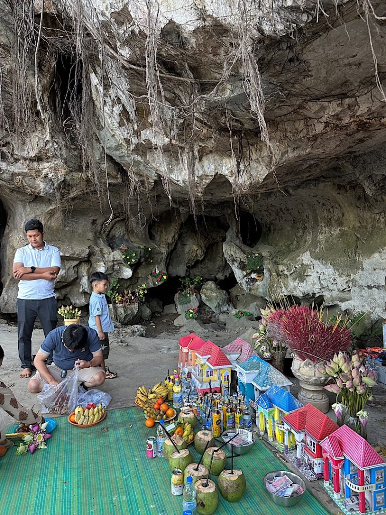 Links steht ein Mann neben einem Altar in einem Höhlentempel. In einer Vase stecken Räucherstäbchen. Davor sind Häuser aus Papier als Opfergaben.