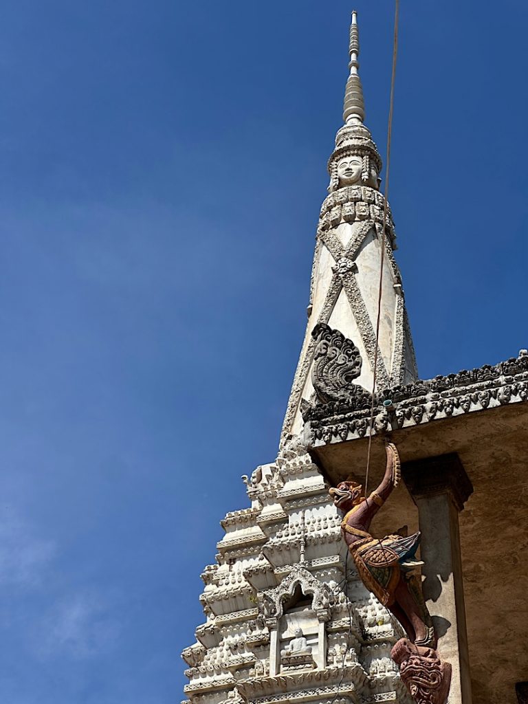 Vor strahlend blauem Himmel ist die Spitze einer Pagode zu sehen. Angeschnitten davor die Ecke eines Tempels. Oben auf der Dachkante ist die Darstellung der Schlange Naga. Darunter hält ein Garuda die Ecke.