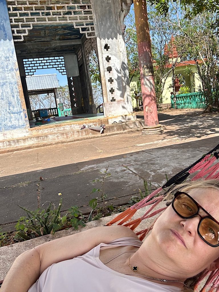 Im Vordergrund sieht man den Kopf und den Arm einer Frau, die in einer Hängematte liegt. Im Hintergrund ist ein Tempel zu sehen.