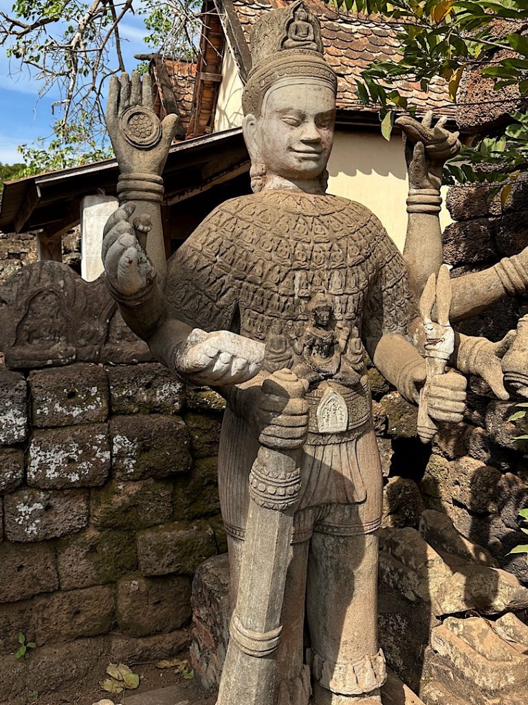 Neben einer Mauer steht eine Figur, die einen Vishnu darstellt. Sie hat 6 Arme. In den Händen hält sie Werkzeug.