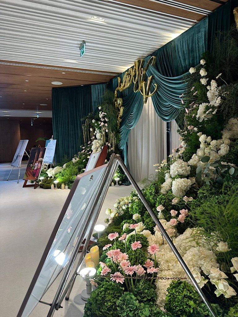 Im Vordergrund steht ein Bildständer. Dahinter ist Blumenschmuck in weiß, grün und rosa. Rechts ist eine Tür mit Grünem und weißem Vorhang geschmückt.