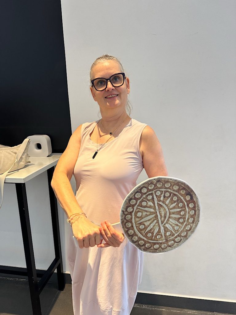 Frau steht vor einer weissen Wand. Links liegt ihre Handtasche auf einem Regal. Sie lacht in die Kamera. Sie trägt ein helles Kleid. Vor sich hält sie ein Schild auf der eine alte Münze abgebildet ist