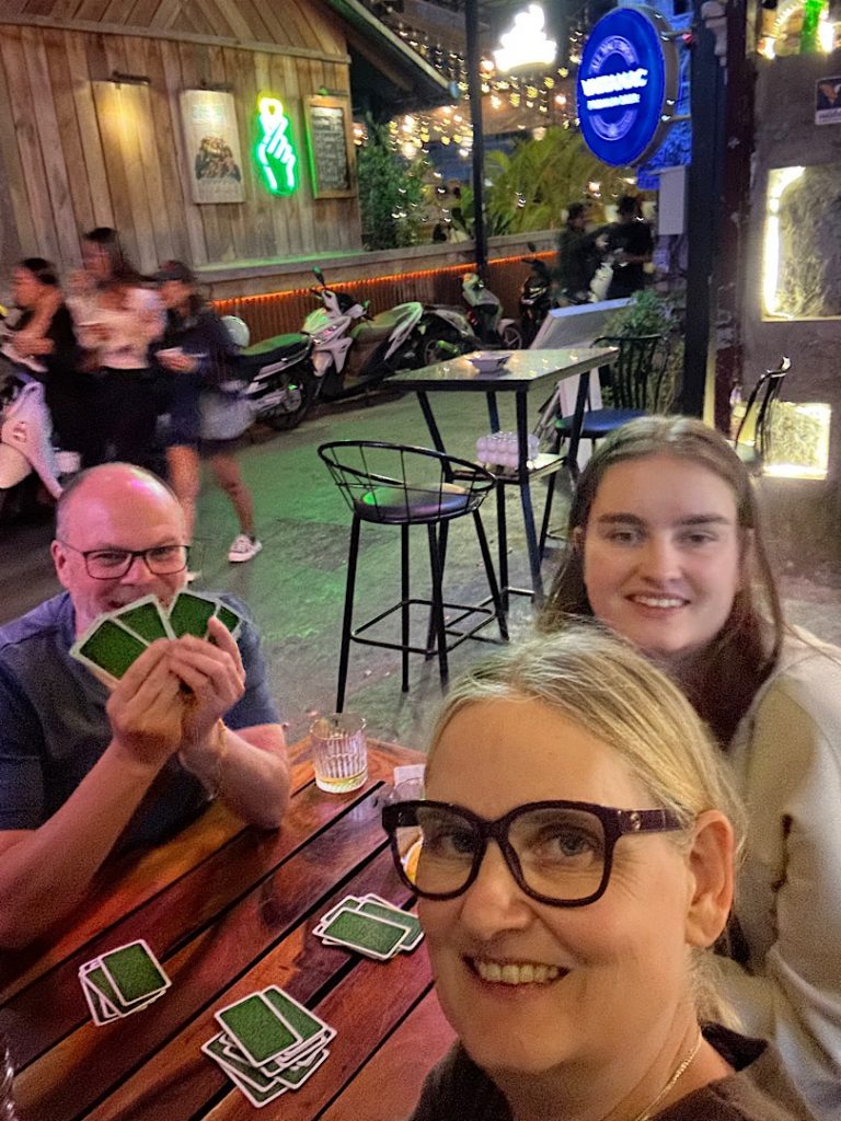 Drei Personen lachen in die Kamera für ein Selfie. Der Mann hat Spielkarten in der Hand. Andere Karten liegen auf dem Tisch.