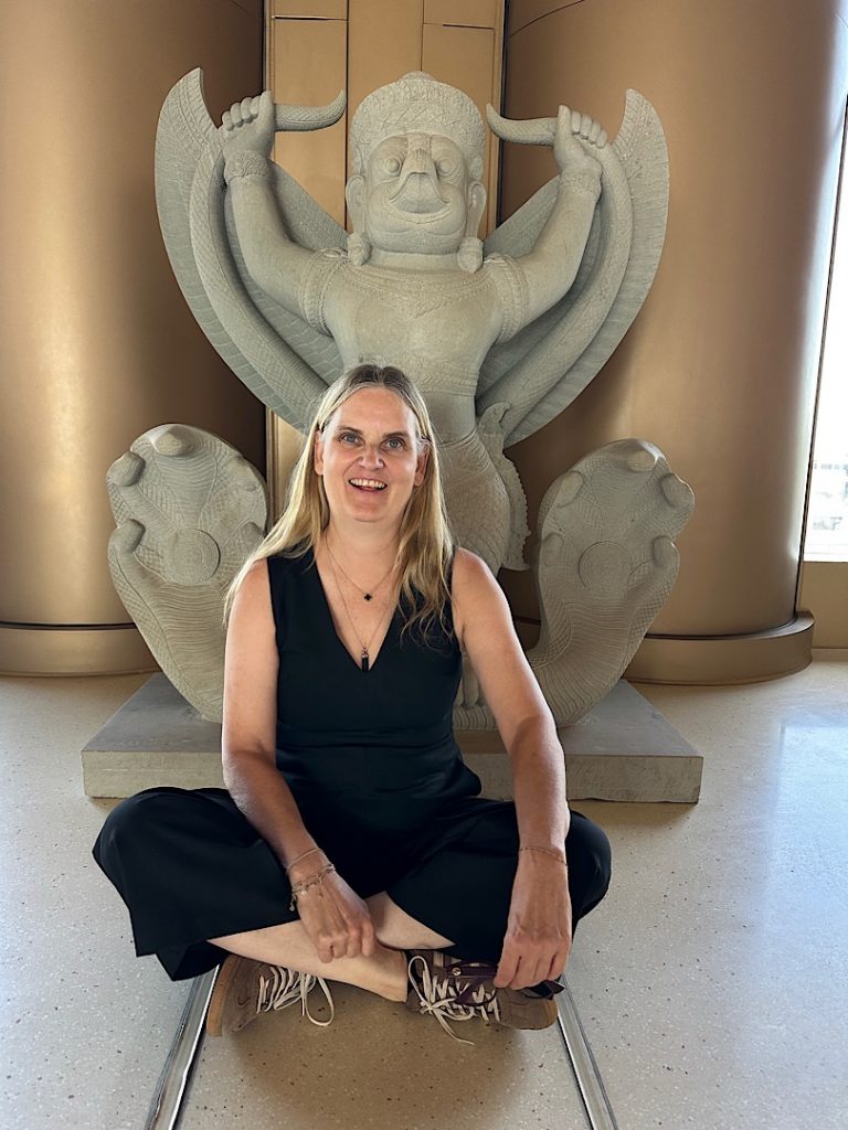 Eine Frau sitzt vor einer steinernen Figur, die Garuda darstellen soll. Sie sitzt im Schneidersitz und lacht in die Kamera. Sie trägt einen schwarzen Hosenanzug.