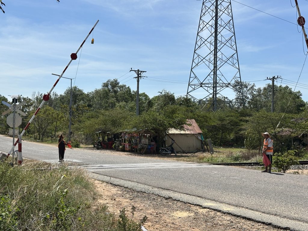 Es ist ein Bahnübergang in Kambodscha zu sehen. Zwei Menschen ziehen an Seilen die Schranke halb herunter. Sie schauen, ob der Zug kommt.