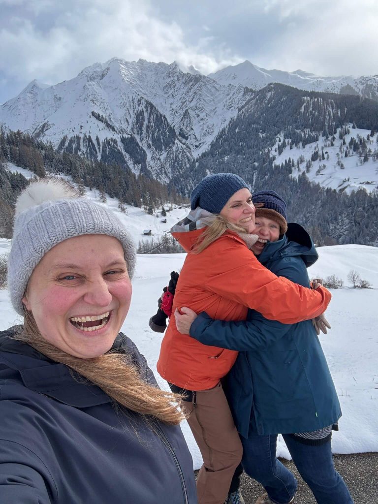 Im Voergrund lacht eine Frau. Sie trägt eine Mütze. Im Hintergrund umarmen sich zwei Frauen. Dahinter schneebedeckte Berge.
